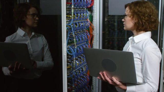Caucasian female IT technician in glasses standing beside opened server rack in data center and testing equipment via laptop computer