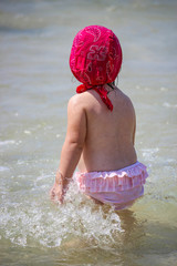A cute and sweet little girl plays with water and sand on the seashore. Child with pink swimsuit and red bandana, back view, walks in the shallow water of the shore. Sea foam and waves.