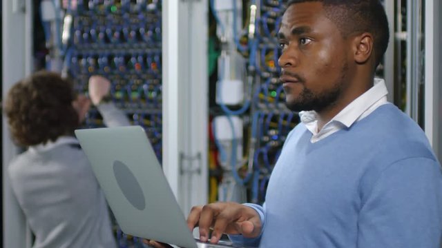 African american IT specialist working on laptop in data center while female technician inspecting server racks in the background