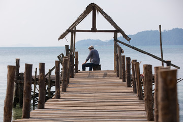 Fototapeta premium Sad old man in a cap sitting on the pier and looking at the sea, the view from the back