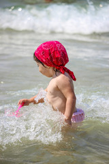 A cute and sweet little girl plays with water and sand on the seashore. Child with pink swimsuit and red bandana, plays with a plastic scoop. Sea foam and waves.