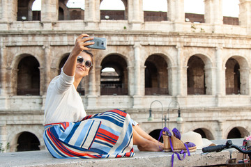 Beautiful young woman taking selfie pictures with smartphone sitting in front of colosseum in Rome at sunset.