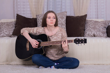 A young woman plays a black acoustic guitar on the carpet in the room.