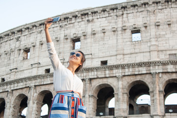 Fototapeta na wymiar Beautiful young woman taking selfie pictures with smartphone standing in front of colosseum in Rome at sunset.