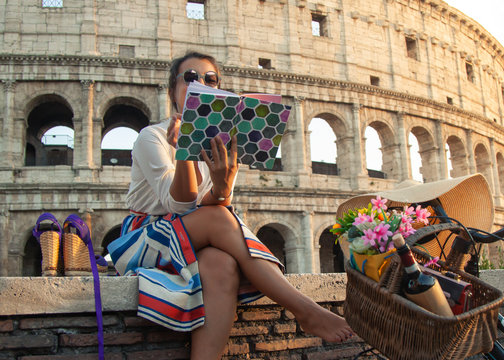 Beautiful young woman reading book sitting in front of colosseum in Rome at sunset.