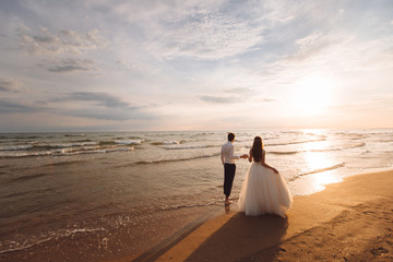Elegant gorgeous bride and groom walking on ocean beach during sunset time. Romantic walk newlyweds...