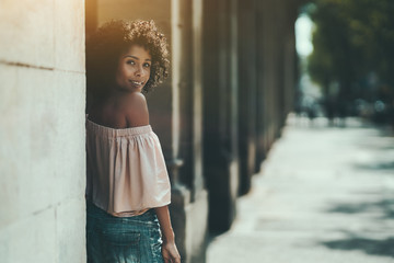 The portrait of a young cute Brazilian woman leaning against the column outdoors; a dazzling smiling African-American female near the stone pillar, with a copy space place for your advertising text