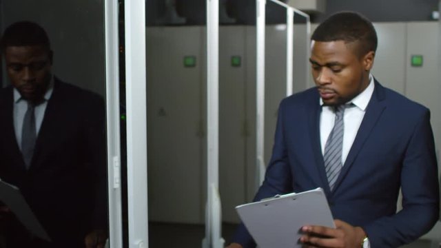 African american IT engineer in formal suit walking along corridor in data center, opening server rack, inspecting optical fiber cables and taking notes