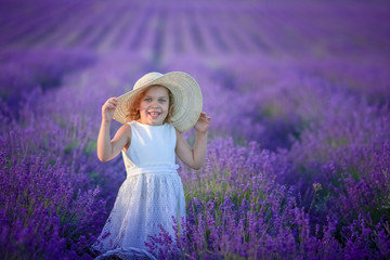 Cute young girl walking on a lavender field in white dress and hat on her head. Cute child face and nice hair with lavender bouquet 