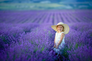 Cute curly young girl standing on a lavender field in white dress and hat with cute face and nice hair with lavender bouquet and smiling.