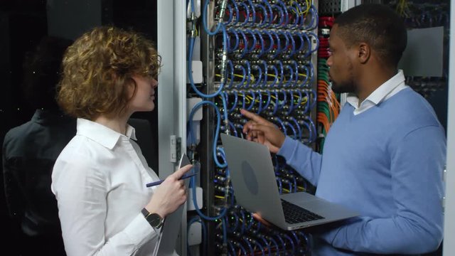 African american IT engineer typing on laptop and explaining something about server rack to cheerful female Caucasian colleague while working together in data center