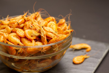 A glass bowl full of delicious orange shrimps on a gray table. Nearby in the background are two cooked shrimps. A plate of shrimp. Snack and appetizer for beer.
