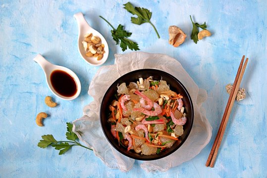Refreshing salad with pomelo, shrimps, carrots and cashew nuts in a black bowl on a light blue background. Asian cuisine. Top view.
