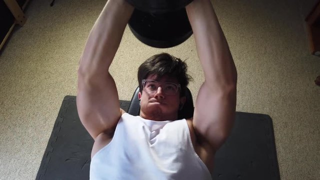 Camera looking straight down on a body builder lifting a heavy dumbbell over his head while lying on a bench.