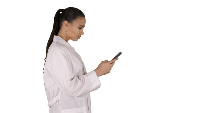 Cheerful cute young woman doctor typing text on phone while walking on white background.