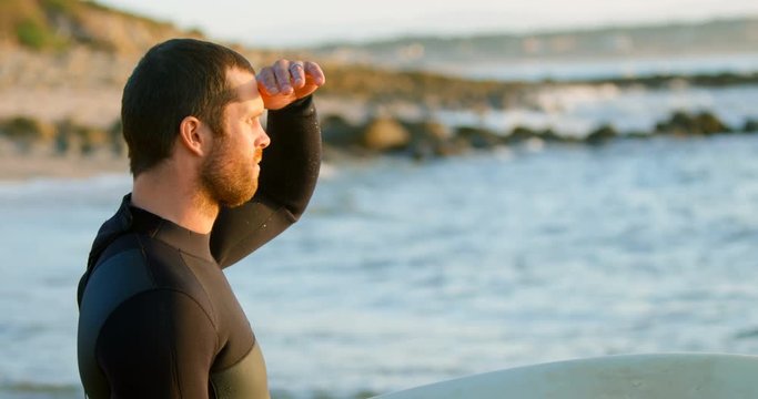 Side view of mid-adult caucasian male surfer with shielding eyes looking at sea at beach 4k