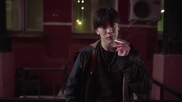 arrogant and confident asian boy is smoking cigarette in evening in courtyard, looking at camera