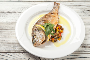 Dorada stuffed with vegetables. White plate with grilled dorada fish on a wooden table close up and copy space