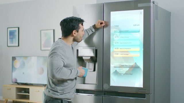Handsome Young Man Walks Over to a Refrigerator While Drinking His Morning Coffee. He is Checking a To Do List on a Smart Fridge at Home. Kitchen is Bright and Cozy.