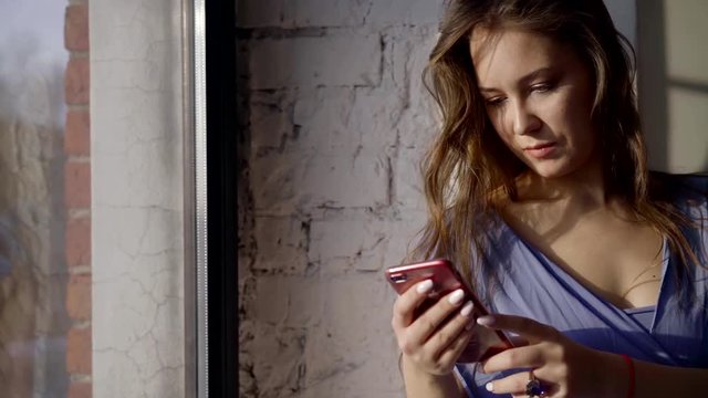 happy brunette girl is viewing funny pictures in her smartphone sitting near window with sunlight