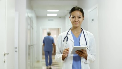 Handheld tracking shot of young female doctor in lab coat and scrubs working on tablet in hospital corridor, then looking at camera and smiling - Powered by Adobe