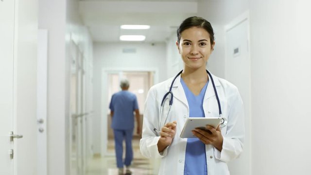Handheld tracking shot of young female doctor in lab coat and scrubs working on tablet in hospital corridor, then looking at camera and smiling