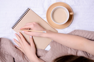 Woman in the white bed cup of coffee in hand notebook pencil brown blanket, top view