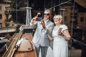 Portrait of smiling senior man and woman sitting in cuddles outside. Mature male is holding mobile and photographing with joy. They are bonding to each other with love and delight