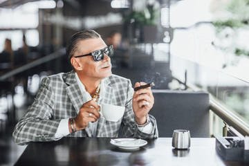Stylish attractive senior in summer terrace chilling with cup of latte and looking forward to
