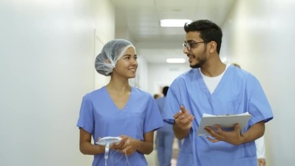 Dolly shot of cheerful female nurse with IV bag walking along hospital corridor and chatting with friendly male colleague writing on clipboard - Powered by Adobe