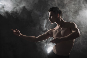 Fototapeta na wymiar A young male ballet dancer with black leggings and a naked torso performs dance moves against a gray grunge background, with a light of lights and smoke.