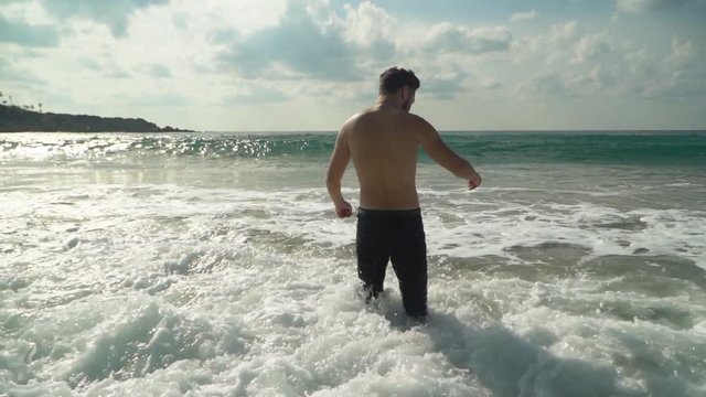 Young man in jeans enters shallow sea water with his back to camera in slow motion. The waves roll on shore. Summer at the beach