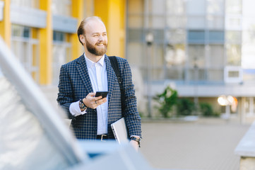 Happy attractive young and bearded businessman walking near business center and using mobile phone while holding laptop.