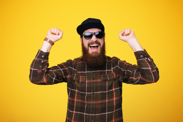 young hipster man isolated over yellow background. Bearded man wearing sunglasses and hat acts like he won something