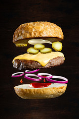 Floating burger isolated on black wooden background. Ingredients of a delicious cheeseburger with ground beef patty, cheese, onions and cucumbers - obrazy, fototapety, plakaty