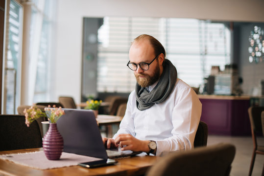 Image of bearded man wearing glasses sitting in cafe while using laptop, freelancer in work.