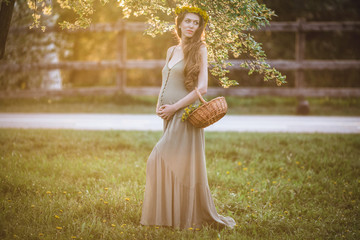 Fototapeta na wymiar Pregnant pretty woman is wearing wreath of yellow dandelion flowers posing over sunset lights , spring time