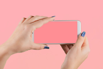 Closeup view of two female hands holding white modern smartphone with blank pink screen isolated on pink background. Fingernails with modern glossy blue manicure. Horizontal color photography.