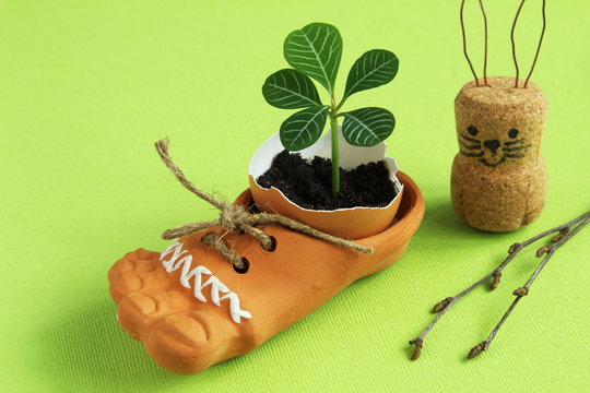 Fragile tender sprout in eggshell in brown ceramic shoe with jute cord, homemade Easter bunny from cork and paper clips and birch twigs on bright green background. Easter concept.