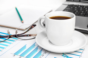Cup of coffee with business graphs and laptop at office workplace.