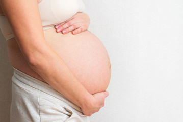 Pregnant woman holds hands on belly on light background. Pregnancy, maternity, preparation and expectation concept. Close-up, copy space, indoors. Beautiful tender mood photo of pregnancy