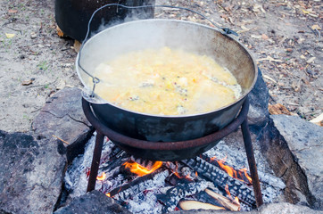 Large metal pot with a lid and handle on tripod used for cooking. Cooking in a bowler hat on the...