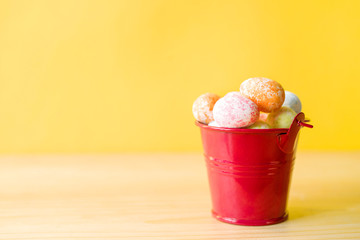 Small red bucket with bright Easter decorative eggs on a yellow background, close up, soft focus. Easter background