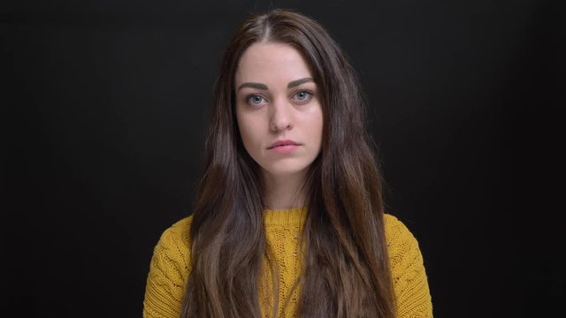 Portrait of young long-haired brunette caucasian girl in yellow sweater watching attentively into camera on black background.