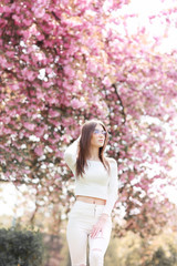 Outdoor portrait of young beautiful happy smiling lady posing near flowering tree. Model wearing stylish accessories clothes. Girl closed her eyes. Female beauty fashion concept. City lifestyle.