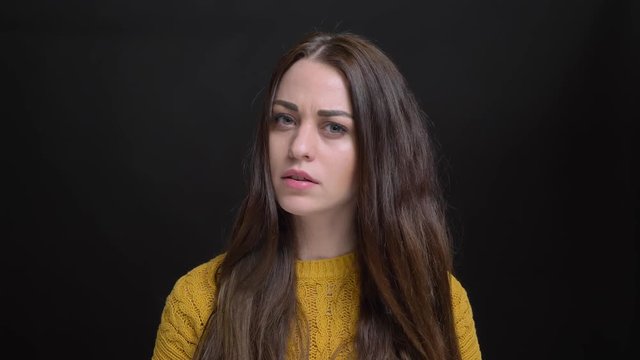 Portrait of young long-haired brunette caucasian girl in yellow sweater watching in confusion into camera on black background.