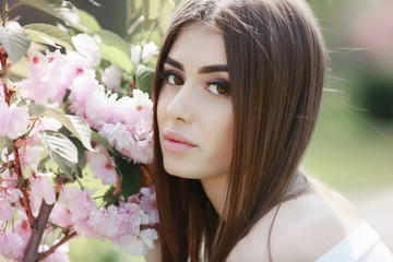 Naklejka premium The beautiful girl with a romantic hairstyle and a professional make-up enjoys a smell of pink colors in a garden. The girl dreams. A portrait of the beautiful girl model in the spring in the park.