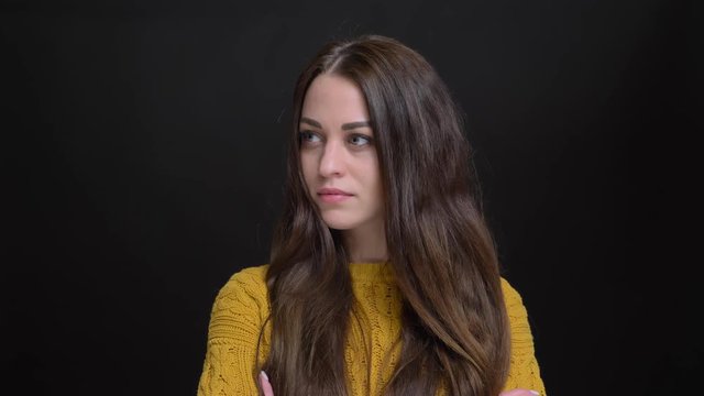 Portrait of long-haired brunette caucasian girl in yellow sweater with crossed arms watching seriously into camera on black background.