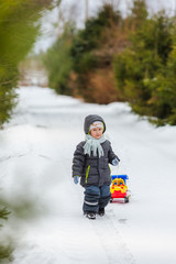 Fototapeta na wymiar Smiling boy walks on snowy street with toy dump trucks