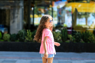 Naklejka premium Fashion kid summer in the city stands on a streetl. Trendy girl in sunglasses standing on the street at sunset.